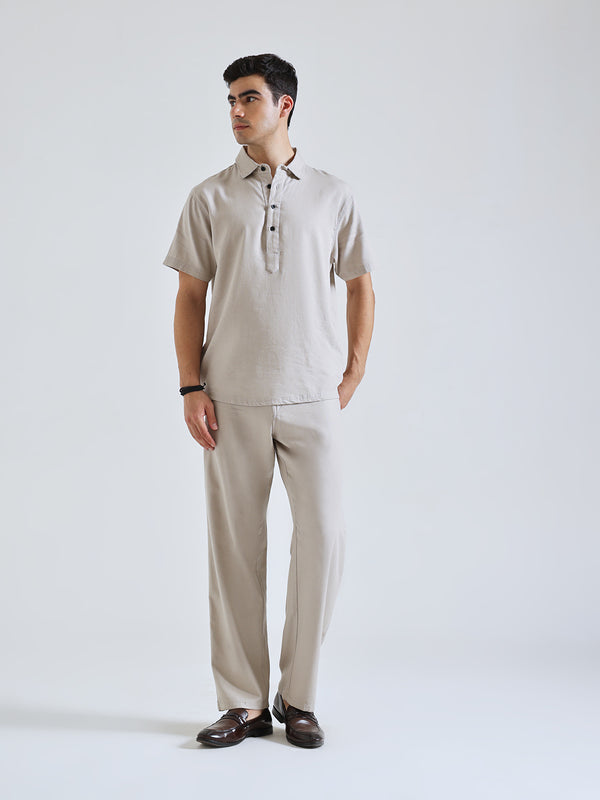 A man modeling a light beige short-sleeve linen polo shirt and matching trousers, paired with dark brown leather loafers, against a plain light gray studio background.