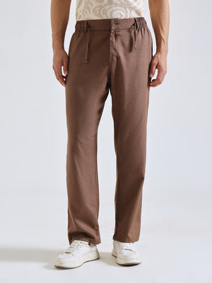 A person wearing brown straight-leg trousers with an elastic waistband and drawstring, paired with white sneakers against a light gray studio background.