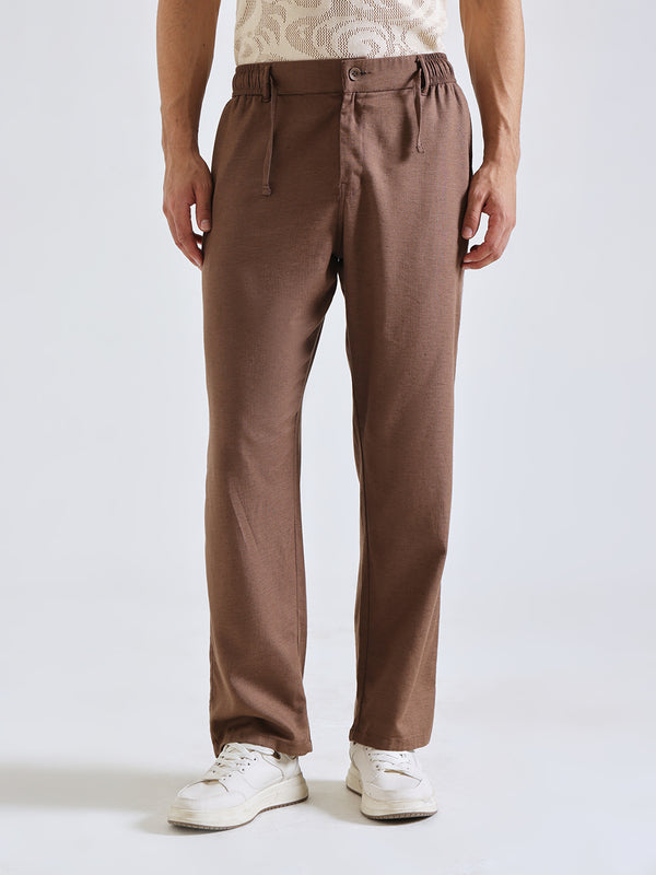 A person wearing brown straight-leg trousers with an elastic waistband and drawstring, paired with white sneakers against a light gray studio background.
