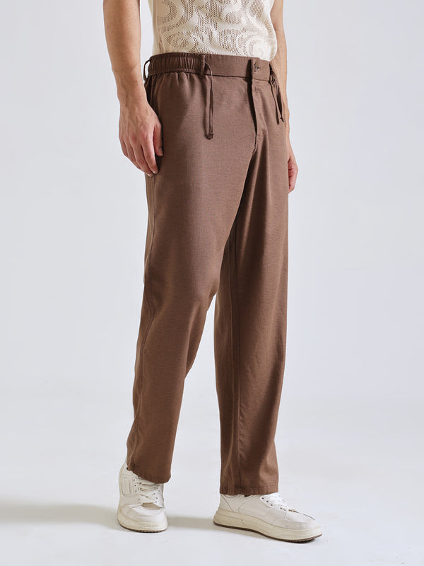 A person wearing brown drawstring trousers, a cream lace top, and white sneakers, photographed against a clean light gray studio background.